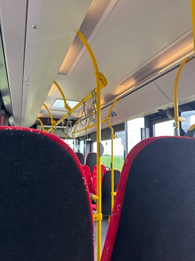 This is an interior shot of a bus, looking forward down the aisle. The seats are upholstered in black fabric with red trim. Yellow support poles and handrails are visible throughout the bus. The ceiling is white with overhead lighting. The windows show a blurry view of a green, grassy landscape. The image appears to have been taken from one of the seats. There are no people visible in the image. The location is given as Breisach am Rhein, Germany.