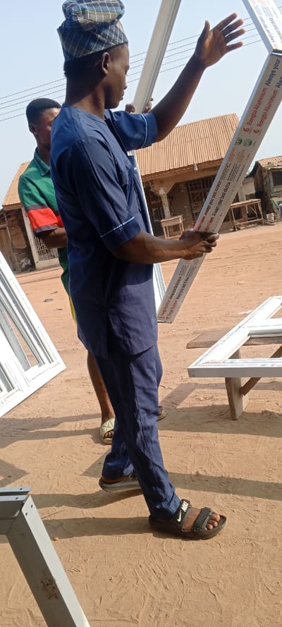 The image depicts an outdoor scene in Ago-Abo, Nigeria, on a dry, light brown dirt ground under direct sunlight. A dark-skinned male, positioned in the foreground, wears a blue and grey plaid cap, a dark blue traditional tunic with light blue trim, dark blue trousers, and black Nike brand sandals. He holds two long, white aluminum profiles vertically. The profiles are labeled "Rongtai Aluminium Always your best choice." His left hand is extended upwards, open towards the top of one profile, while his right hand grips the lower sections of both. A second dark-skinned male is partially visible behind him, wearing a green polo shirt with red and black stripes on the sleeves and light green slide sandals. In the mid-ground, multiple white window frames or frame components are visible on the ground and resting on a crude wooden workbench. The background features simple residential or workshop structures with reddish-brown corrugated metal roofs and overhead power lines. The scene indicates an active site for window or door frame fabrication or assembly.