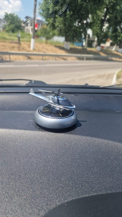 An eye-level, medium shot captures a solar-powered helicopter model on a car's dashboard. The dashboard is dark gray and textured. The helicopter, with a silver body and black accents, rests on a circular gray base that features a solar panel. The background, visible through the car's windshield, shows a blurred scene of a road, a guardrail, trees, houses, and a utility pole. The sky is bright blue with some clouds. The image appears to have been taken in Cricova, Moldova.
