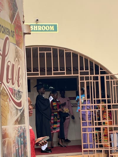 The scene depicts a gathering at the arched entrance of a building in Madina, Ghana. A cream-colored wall forms the background, featuring a green sign with partially visible white text reading "SHROOM" (likely "WASHROOM") mounted above the arch. A small white security camera is affixed to the wall above the sign. The arched entrance is secured by an ornate light-colored metal gate with vertical and horizontal bars. A red carpet or rug is visible on the floor just inside the entrance.
In the left foreground, the partial profile of an adult individual with dark skin, wearing a red garment, is visible, holding a dark rectangular object in their right hand.
Inside the entrance, a young man, presumably a graduate, stands facing right, dressed in a black graduation gown and cap, white trousers, and black shoes; he appears to be adjusting his left shoulder. Adjacent to him, a young woman, also dressed for graduation, wears a dark top, dark trousers, and black high-heeled shoes, with a colorful black, red, yellow, and green Kente-patterned stole draped over her left shoulder. She holds a small, cylindrical woven basket-like object with a handle. Behind these two, an adult man in a light brown long-sleeved shirt and dark trousers stands observing the scene.
Further right, behind the metal gate, a woman in a purple patterned traditional dress is visible, holding a baby dressed in a light blue garment. Another person in colorful Kente-patterned attire is partially visible beyond them. Indistinct figures are present in the deeper background. The individuals are gathered at the doorway, indicating an event or transition point.