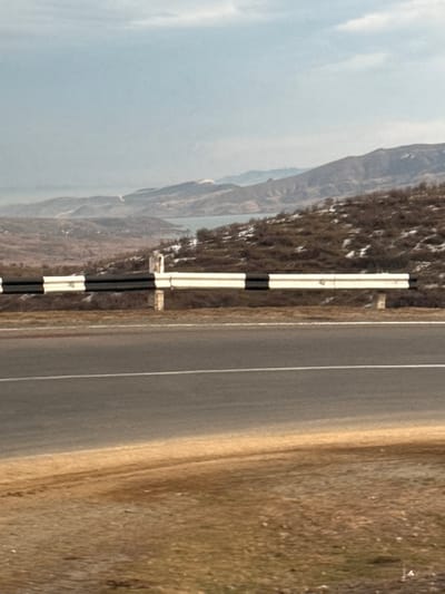 The image displays a desolate, mountainous landscape in the vicinity of Voskepar, Armenia. In the foreground, a two-lane asphalt road with a solid white center line runs horizontally. To the right of the road is a narrow dirt shoulder, beyond which a metal guardrail, painted with alternating black and white stripes, is erected on several light-colored concrete posts. The terrain immediately behind the guardrail consists of dry, brown vegetation and sparsely distributed patches of snow, sloping upwards towards the midground. A body of water, likely a lake or reservoir, is visible in the middle distance, nestled among rolling hills. In the far background, multiple layers of hazy mountains extend under a light blue sky with scattered white clouds. No people, vehicles, or active interactions are present within the frame.