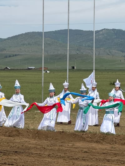 In Orongoi, Russia, a group of women in traditional white and blue attire, complete with pointed hats, perform a dance in a grassy field. They are holding colorful silk scarves, red, white, green, and blue, which they manipulate in unison. The background features rolling hills covered in green vegetation and scattered trees, with several tall poles, one bearing a white flag with red markings, standing in the middle ground. In the distance, a few wooden buildings and a white vehicle are visible. The foreground consists of a patch of brown, disturbed earth.