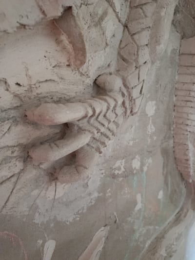 This is a close-up, dimly lit, horizontal photograph of a textured, light-colored, stucco or plaster sculpture. The sculpture appears to be part of a larger, unfinished artwork, possibly a mural or a decorative relief. The texture of the stucco is rough and uneven, with visible imperfections and some chipping.

The primary subject in the foreground is a segment of a sculpted limb, likely a leg, from a human or humanoid figure. The limb is depicted with a segmented texture resembling armor or scaled skin, with a zigzag pattern etched into its surface. A portion of the leg appears to be bent, with details suggesting knees or joints. To the left of this limb, two distinct, rounded forms can be seen, which could represent feet or hands, also rendered in a simplified, stylized manner with visible segmentation.

The background is also sculpted and textured, with areas of rough plaster and what appear to be deliberate incised lines creating patterns that could represent bricks, stones, or scales. Faint traces of color, possibly from chalk or pastel, are visible on the surface, including pinkish and white marks, suggesting it may be an interactive or outdoor art piece. Some areas of the stucco show evidence of being wet or stained, with faint green and white streaks running down the surface.

The overall impression is of an unfinished artistic project, possibly created in an outdoor setting given the texture and potential for weathering. The lighting is subdued, suggesting it might be daytime with overcast conditions or artificial lighting. No text is visible in the image. The location context is Smarhon, Belarus.