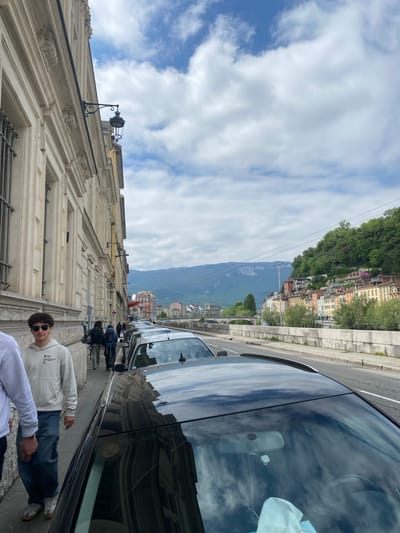 Here's a description of the image:

The image presents a street scene. The photo is taken from the sidewalk, facing a street lined with vehicles. On the left is a building with ornate architectural details. People are walking on the sidewalk. In the background, across the street, is a hillside with buildings. Mountains are visible in the distance. The sky is cloudy. The location appears to be in a European city. Grenoble, France is the city.