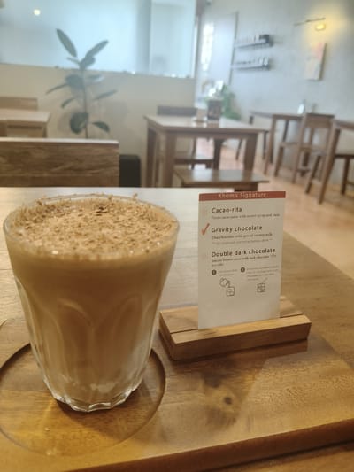 The image shows a close-up of a glass of iced chocolate drink topped with chocolate shavings. The drink is served on a wooden tray. To the right of the drink is a small wooden stand holding a menu card. The menu card is titled "Khom's Signature" and lists three drink options: "Cacao-rita," "Gravity chocolate," and "Double dark chocolate." The "Gravity chocolate" option is checked with a red tick mark, indicating it is the selected drink. The background of the image shows the interior of a cafe with wooden tables and chairs. There is a plant on a shelf in the background. The lighting suggests it is daytime, and the overall ambiance is relaxed and inviting. The cafe appears to be located in Chiang Rai City Municipality, Thailand, given the context of the menu and the style of the cafe.