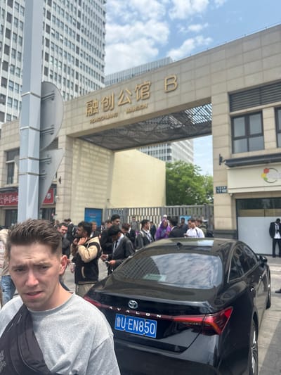 The image captures a man taking a selfie. Behind him is a crowd of people gathered in front of a building with the Chinese characters 融创公馆 (Rongchuang Mansion) and the English translation "RONGCHUANG MANSION" on the gate. A black car, with license plate U·EN850, is parked nearby. The building in the background has a street address sign that reads "1746". Given the characters 融创公馆, this appears to be a residential complex in Huangdao District, Qingdao, China.