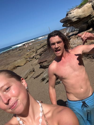 The image displays two individuals on a rocky beach at Coalcliff, Australia, under a clear blue sky. In the lower-left foreground, a female with wet, slicked-back hair is positioned, wearing a patterned bikini top and a white beaded necklace, with her face directed towards the camera exhibiting a slight smile. To her right and slightly behind her stands a shirtless male with wet, medium-length dark hair. He wears blue swim shorts, has a visibly sunburned chest, an open mouth, and his right hand is raised, performing a "shaka" gesture. The background reveals an extensive rocky intertidal platform stretching towards the ocean, with waves visible breaking in the distance. Several indistinct human figures are present further along the distant shoreline. A prominent, layered rock formation is visible on the far right. The scene is brightly illuminated by direct sunlight.