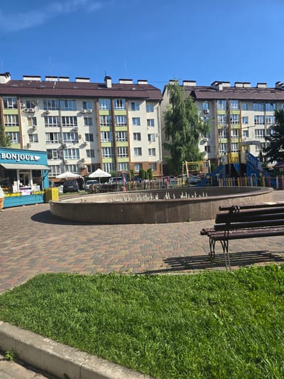 In Petropavlivska Borshchahivka, Ukraine, a residential courtyard is depicted under a clear blue sky. The scene features modern apartment buildings with beige and green facades, featuring numerous windows and air conditioning units. In the foreground, a circular stone fountain with a series of water spouts is centrally located. To the left of the fountain is a light blue kiosk with the word "BONJOUR" prominently displayed, suggesting a small business, possibly selling ice cream or refreshments. Adjacent to the kiosk, a patch of vibrant green grass meets a decorative paved walkway constructed from interlocking bricks in shades of red, yellow, and gray. A dark wooden park bench with wrought iron legs is positioned on the paved area, facing the fountain. Behind the fountain and to the right, a colorful children's playground is visible, complete with slides, climbing structures, and a protective fence. Several cars are parked near the buildings and playground. The overall environment is tidy and well-maintained, indicative of a residential area designed for communal use and recreation.