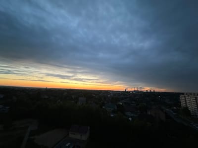 The image depicts a twilight scene over a suburban area, likely in the vicinity of Шчомысліцкі сельскі Савет, Belarus. The sky is a dramatic canvas of dark gray clouds overhead, gradually giving way to a warm orange and yellow gradient at the horizon, indicating either sunrise or sunset. Silhouetted against the horizon are several tall, industrial chimneys emitting plumes of smoke or steam, suggesting the presence of a power plant or factory in the distance.
Below the sky, the landscape transitions into a dark silhouette of a residential area. Numerous houses with pitched roofs are visible, interspersed with dense patches of trees. In the foreground, a few buildings are more clearly discernible in the fading light. One is a small, light-colored structure with a darker roof, possibly a shed or a small utility building. To the right, a multi-story apartment building with numerous windows stands out, its facade illuminated by the last rays of light. A road or pathway runs alongside this building, with several parked cars visible. The overall impression is one of a quiet, encroaching evening in a semi-urban environment. There are no visible people in the image.