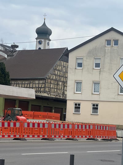 This image captures a street scene in Krauchenwies, Germany, under an overcast sky, focusing on a construction site in the foreground against a backdrop of historic and residential buildings. In the upper center, a prominent church tower with a dark green, bulbous dome topped by a cross and a golden sphere features a clock face displaying approximately 1:30 or 2:30. To the left of the church, a large stork nest is visible on a platform extending above a dark brown tiled roof. Below the tiled roof, an aged half-timbered structure is present, situated above a lower building with multiple dark green garage doors.
To the right of these structures stands a multi-story residential building with a light beige stucco facade, featuring several rectangular windows with white frames; some windows display white roller shutters, while others show wavy-patterned curtains or blinds. Weathering stains are visible on the facade. A yellow, diamond-shaped priority road sign is partially visible on the far right.
In the mid-ground, a construction area is delineated by a continuous line of orange and white striped temporary barriers. Behind these barriers, a red compact construction vehicle, specifically a Komatsu mini skid steer branded "STRINGEL," is parked. To the left of the vehicle, a grey concrete structure or overhang is visible. The foreground features a dark asphalt road with a single white lane marking and two dark bollards on the right. No people are visible in the scene.
