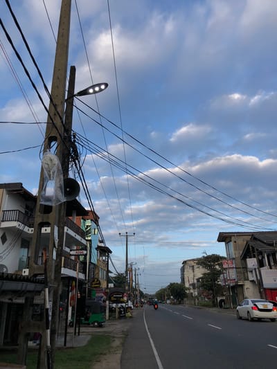 This media file depicts a street scene in Habaraduwa, Sri Lanka. The image is oriented vertically, with the sky taking up a significant portion of the frame. The sky is filled with dramatic, overcast clouds, suggesting it might be late afternoon or early evening, or a cloudy day.
The foreground is dominated by a utility pole, heavily laden with electrical wires. A street light with three lamps is attached to the pole, emitting a bright light. Below the street light, a large, dark speaker is mounted, covered in a clear plastic bag.
The midground reveals a street lined with buildings. These appear to be shops and residences, with multiple stories and balconies. Various signs are visible, some with text in a language that is likely Sinhala, given the location. One sign indicates "ROOMS," suggesting accommodation. A white car is parked on the side of the road near one of the buildings. Further down the street, a green tuk-tuk is visible, along with traffic signals showing red lights, indicating a stopped flow of traffic.
The background shows more of the street and buildings, stretching into the distance. The road itself is paved and appears to be a main thoroughfare. The overall scene conveys a sense of a bustling, everyday street in a town in Sri Lanka, with the weather adding a slightly dramatic atmosphere. There are no people clearly visible in the frame, though their presence is implied by the activity on the street.