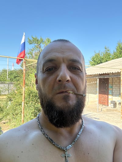 A man with a beard and a bald head is shown from the chest up, facing forward. He has a cigarette in his mouth and is wearing a silver chain with a cross pendant. In the background, a Russian flag flies from a pole, and a brick building with a brown door is visible. The scene appears to be outdoors, with greenery and a clear blue sky. The man's bare shoulders are visible. This scene is located in the city of Zhirnov, Russia.