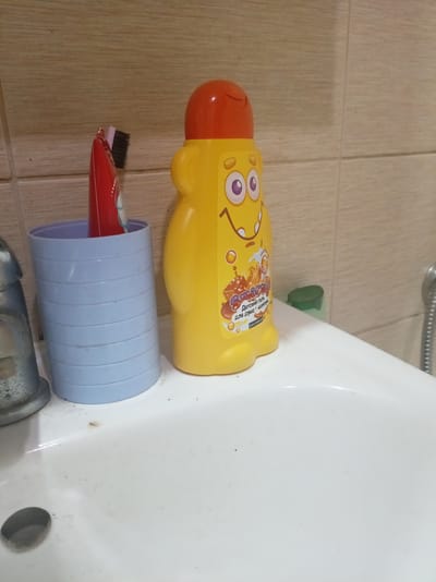 The image shows a bathroom sink with several items on its surface. A blue ridged cup holds a red toothbrush. Next to the cup is a bright yellow bottle of "Smashy" children's gel for bath and shampoo, featuring a cartoon character face and text in Russian that translates to "Children's gel for bath + shampoo." The bottle has an orange cap. In the background, light brown tiled walls are visible. A silver faucet is partially visible on the left side of the sink, and a portion of a silver shower hose is seen on the right. The sink itself is white and appears to be in use, with some water residue and small specks of dirt. The overall scene suggests a typical bathroom setting, likely in a residential location in Kamensk-Uralsky, Russia. There is no clear indication of the time of day or weather.