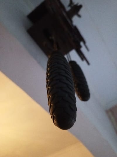 An interior scene in Keo Kouang, Laos, is captured from a low angle looking upwards towards a ceiling. The primary subjects are two dark, pendulous objects, likely weights from a cuckoo clock, suspended from an out-of-focus, dark wooden mechanism partially visible at the top. The foreground object is a sharply focused, elongated, pinecone-shaped weight, exhibiting a distinct textured, scaly surface in a dark, possibly black or dark brown, color. A second, smaller, and less defined pinecone-shaped weight is visible behind and to the right of the prominent one. The ceiling appears white or light grey, with a warm, yellow-orange light source illuminating the lower-left portion of the frame, creating a glow on the ceiling and an adjacent vertical surface. The overall lighting is dim, with the upper section of the image appearing darker. No individuals, active events, or explicit interactions are depicted.