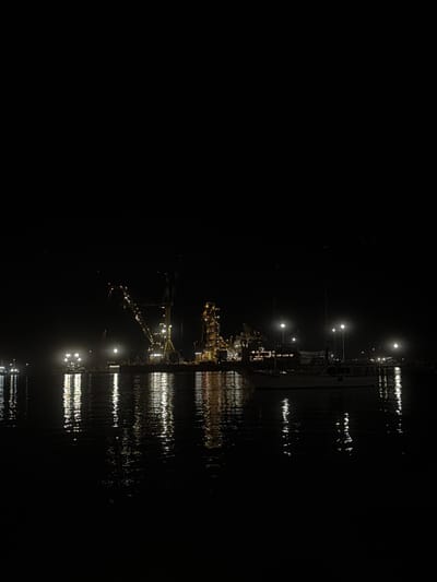A nighttime scene displays a pier or dock extending into water. Lights illuminate the structure, reflecting in the water below, creating shimmering patterns. The darkness envelops the majority of the scene, obscuring details beyond the lit area. Distant lights suggest a cityscape or industrial complex at the pier's end. The overall impression is one of solitude and mystery.