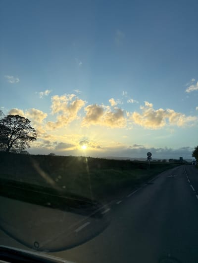 The image captures a rural roadside scene near Ruyton-XI-Towns, United Kingdom, likely viewed from inside a vehicle. The sun is positioned low on the horizon, shining through scattered clouds, creating a golden, hazy light. A road runs along the right side of the frame, adjacent to a grassy verge and foliage. On the left, a silhouette of a tree is visible against the sky. A road sign is also seen on the right side. The view is somewhat obscured by the interior of the car.