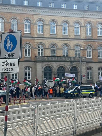 The image depicts a protest occurring in front of a building in Osnabrück, Germany. A crowd of people is assembled, many holding Palestinian flags. Some individuals are carrying signs, including one stating "Völkermord in Gaza, Deutschland liefert Waffen" (Genocide in Gaza, Germany delivers weapons). A police van is present, and a police officer is nearby with a bicycle. White plastic barriers line the street. A street sign is visible, designating a pedestrian zone and indicating bicycle access is permitted between 20:00 and 10:30. The buildings feature arched windows and brick facades.