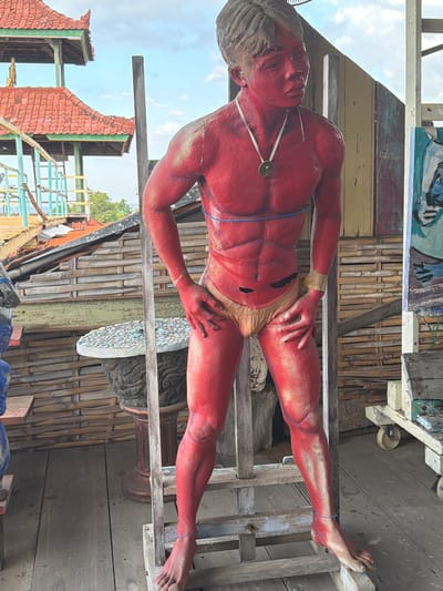 The image depicts a full-body statue of a male figure painted entirely red, standing on a wooden platform. The statue has well-defined musculature, including visible abs, and is wearing a light-colored loincloth. A blue cord is wrapped around the statue's torso, and a thin, lighter cord is around its ankles. A necklace with a circular pendant hangs around the statue's neck. The background reveals a rustic environment with bamboo walls and wooden structures. In the distance, a traditional Balinese-style building with a red tile roof and green trim is visible. To the right of the statue, a wheeled cart with various items, possibly art supplies or tools, can be seen. The overall setting suggests an outdoor artistic or cultural space, potentially a workshop or exhibition area, located in Tejakula, Indonesia. The lighting indicates it is daytime with a partially cloudy sky.