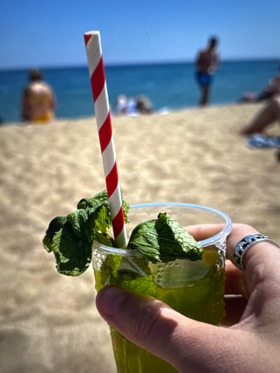 In the foreground, a person's hand, adorned with a silver ring, holds a clear plastic cup filled with a green beverage. The drink is garnished with fresh mint leaves and contains ice cubes. A red and white striped straw protrudes from the cup. The background depicts a sandy beach with the blue ocean and clear sky beyond. Several blurred figures are visible on the beach and in the water, suggesting a summer day at a seaside location. The image does not depict any elements specific to the city of Porrentruy, Switzerland, as it is an outdoor beach scene.
