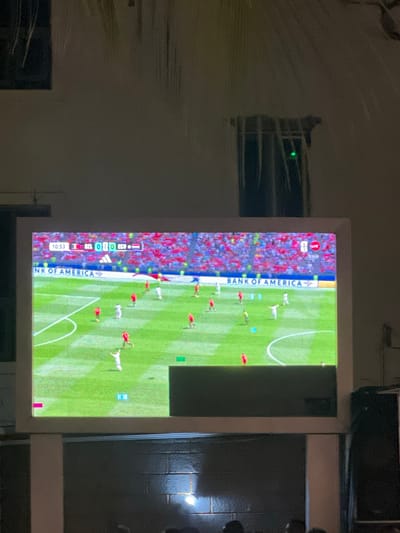 The image shows a large screen displaying a soccer game. The screen is displaying a game between Belgium (BEL) and Egypt (EGY) with the score at 0-0 in the 10:53 minute. The screen is mounted on a dark wall, possibly in a public space or a large room. The scene appears to be in Oba, Nigeria, as indicated by the question. Several people are visible in the foreground, blurred and out of focus, appearing to be watching the game. There are also speakers on the right side of the image. The overall environment is dimly lit.