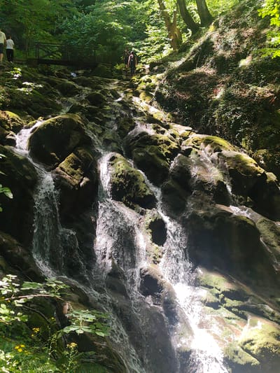 In the city of Teteven, Bulgaria, a group of people is hiking near a cascading waterfall. The waterfall flows over moss-covered rocks, surrounded by lush green foliage and trees. The sun shines through the canopy, creating dappled light on the water and rocks. The people are walking along a path that follows the course of the waterfall. The scene depicts a natural landscape and outdoor recreational activity.