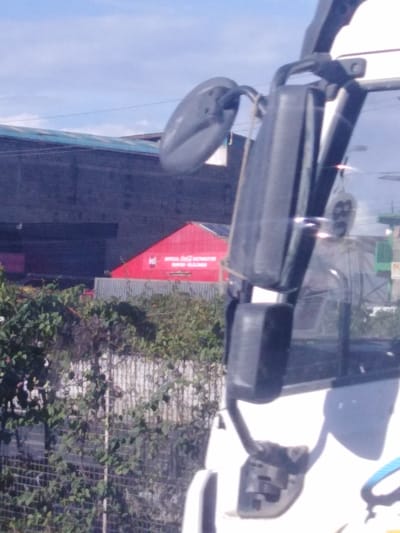A daytime shot depicts a vehicle's side mirror obscuring parts of a cityscape. Behind the mirror, lush foliage conceals a white fence. Further back, a red building, labeled "Official Coca-Cola Distributor," stands prominently near a gray industrial structure under a bright sky. The mirror reflects the exterior environment, adding depth to the image and blending the foreground with the backdrop.