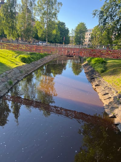 The image depicts a canal with a decorative red metal bridge spanning across it. The canal banks are lined with stone walls and grass. Trees and buildings are visible on either side of the canal, with a clear blue sky overhead. The water reflects the bridge, trees, and sky. The scene appears to be a public space within the city of Oulu, Finland, likely in a park or urban area.