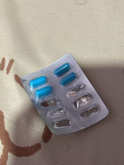A blister pack containing four blue pills lies on a surface patterned with a camouflage design. The blister pack is mostly empty, with only the four pills remaining in their individual compartments. The remaining compartments are open and empty, showing the clear plastic packaging. The pills are cylindrical and opaque blue. The camouflage pattern on the surface beneath the blister pack consists of irregular brown and light tan shapes. The lighting suggests an indoor environment, possibly with artificial light, and there are no discernible indicators of the time of day or weather conditions. No text is visible in the image. The overall scene is simple, focusing on the medication and its packaging against a textured background. There are no people present in the image.