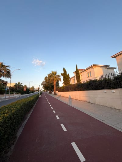 This media file depicts a street scene in Pervolia, Cyprus, during what appears to be late afternoon or early evening, given the warm, golden light cast on the buildings and the long shadows. The image is oriented vertically, showing a clear blue sky with a few wispy clouds on the left side.
The main subjects are urban infrastructure and residential architecture. On the left, a modern building with white walls and a tiled roof is visible, partially obscured by a tall, black wrought-iron fence and lush green bushes. Several cypress trees stand tall along the property line, adding to the Mediterranean feel.
To the right, a wide, reddish-brown paved pathway, likely a bike lane or pedestrian walkway, dominates the foreground. It is marked with white dashed lines, indicating a designated route. This pathway runs alongside a neatly trimmed green hedge. Further down the path, streetlights and traffic signs are visible. One sign is circular with a red border and the number "60" inside, likely indicating a speed limit. Another sign appears to be a blue directional sign.
The setting is a well-maintained residential or resort area. The overall mood is peaceful and tranquil, characteristic of a quiet street in a warm climate at the end of the day. The presence of palm trees and the architectural style of the buildings suggest a coastal Mediterranean location. No people are visible in the image.
