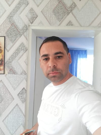 The image shows a man standing in what appears to be a doorway or hallway, with patterned wallpaper on the left and a bright opening to another room on the right. The man is looking towards the camera and is wearing a white t-shirt with a graphic design. His right hand is visible holding a light-colored object with a blue tip. The background on the right shows blue curtains and a bright light source, suggesting a window or another room. The patterned wallpaper on the left features geometric shapes in white, grey, and silver tones. The overall scene appears to be indoors, likely in a residential setting. The location in Bukovlak, Bulgaria is not explicitly indicated in the image.