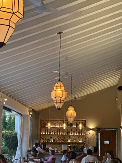 The image shows the interior of a restaurant with a distinctive slatted ceiling. Hanging from the ceiling are several ornate, lantern-style light fixtures. A bar area is visible in the background, stocked with various bottles. Seated patrons can be seen enjoying their meals, creating a bustling atmosphere. Outside, there is a glimpse of lush greenery through a draped opening.