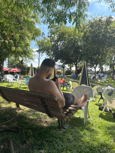 A man with short dark hair and an earring sits shirtless on a wooden park bench in Beit Yitzhak-Sha'ar Hefer, Israel, looking at his phone. He is wearing dark shorts and has white earbuds in his ears. His bare feet are visible. The bench is on a grassy area surrounded by trees. In the background, there are numerous white and blue chairs, some with towels draped over them, and several large, conical umbrellas. Red and white umbrellas are also visible. The sky is blue with some clouds. The scene appears to be a public outdoor area, possibly a park or a recreational facility.
