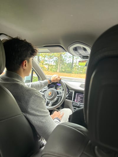 An image taken from the rear passenger seat depicts the interior of a Porsche Macan in Wellesley, United States. A male driver, visible from the back and left side, is at the wheel. He has dark, curly hair and is wearing a light gray turtleneck sweater and light-colored pants. A dark-banded smartwatch is visible on his left wrist. His hands are positioned on the multi-function steering wheel, which features the Porsche crest. The dashboard's infotainment screen shows a menu. Through the windshield, a roadside environment with trees displaying green and autumnal foliage is visible. A dark headrest or partial figure is present in the front passenger seat.
