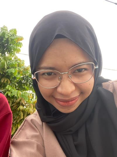 The media file is a selfie of a young woman wearing a black hijab and silver-rimmed glasses. She is smiling and looking directly at the camera. Her face is well-lit, suggesting it is daytime. She is wearing a light brown garment over a red garment, possibly a sweater or jacket. Lush green foliage is visible in the background, along with a bright, overcast sky. The image appears to have been taken outdoors, possibly in a garden or park. The overall mood is cheerful and relaxed. There is no visible text in the image. The location context is Air Molek, Indonesia.