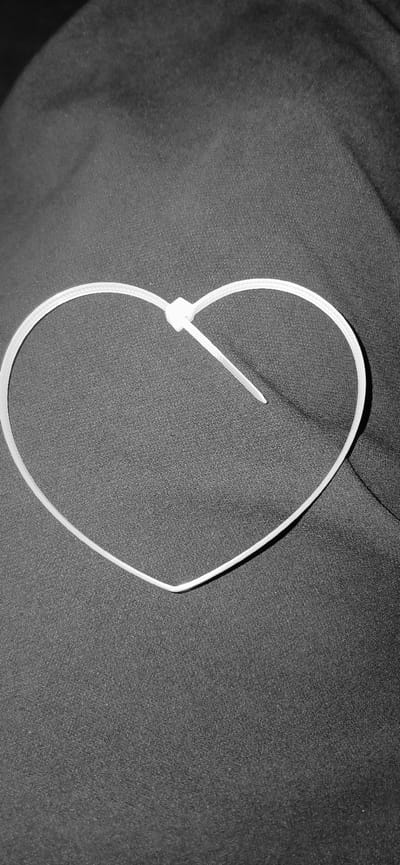The image shows a close-up, black and white view of a white cable tie lying on a dark, textured surface, possibly fabric. The cable tie is partially formed into a heart shape, with the locking mechanism clearly visible in the center. The tail of the cable tie extends upwards and to the right. The textured surface appears to be wrinkled or folded, creating subtle shadows and highlights that add depth to the image. The lighting is soft, and there are no indications of time of day, weather, or specific location cues within the frame itself, beyond the implied indoor setting suggested by the lighting and the nature of the object and background. No text is visible in the image. The overall impression is one of simplicity and focus on the texture of the background and the form of the cable tie.