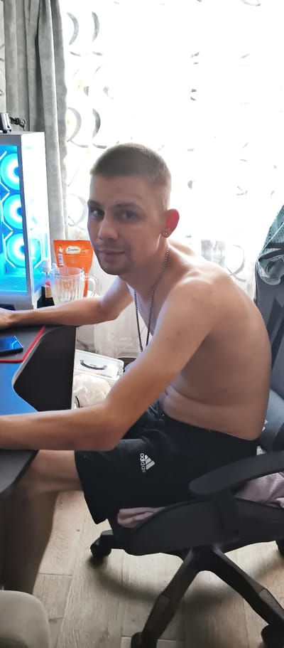 The image depicts a shirtless young man sitting in a black office chair in what appears to be a room with wood-laminate flooring and light-colored curtains. He has short, light brown hair and is wearing black athletic shorts with a white Adidas logo. A silver chain necklace is visible around his neck, and he has a gold earring in his left ear. The man is looking towards the viewer with a neutral expression. To his left, a computer tower with blue-lit fans is visible, along with a desk and a glass mug containing a beverage. An orange packet of "Dreamers" snacks is also on the desk. The overall setting suggests a casual indoor environment, possibly a home office or gaming setup, in Kondrovo, Russia.