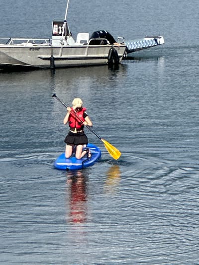 A person is paddleboarding on a body of water with a boat in the background. The person, with blonde hair, is wearing a red life vest over a black top and a black skirt. They are kneeling on a blue paddleboard and holding a paddle with a yellow blade. The boat in the background is silver and white, with a number "33" visible on a black sign. The water is calm with ripples, and the sky appears overcast or hazy. The scene is set outdoors, likely on a lake or calm sea. Without further context or landmarks, the specific location within North Vancouver, Canada, cannot be determined from the image alone.