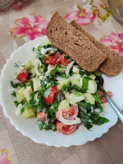 A close-up, overhead shot shows a white, scalloped-edge plate filled with a fresh salad. The salad consists of chopped cucumbers, tomatoes, white onion slices, and dark green leafy herbs, all dressed in a creamy white sauce. Two slices of dark, whole-grain bread are placed on top of the salad, slightly overlapping. A silver fork rests on the right side of the plate, partially submerged in the salad. The plate sits on a tablecloth with a pattern of pink and gold flowers against a neutral background. The scene is brightly lit, highlighting the textures and colors of the food. The overall impression is of a simple, home-cooked meal. The location is in the city of Chernogorsk, Russia.