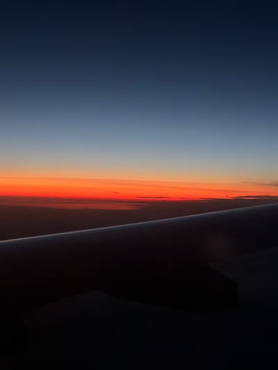 An aerial view captured from an airplane window shows a twilight sky over a dark landscape. The sky transitions from a deep blue at the top to vibrant orange and red hues near the horizon. The silhouette of an airplane wing occupies the foreground, its dark shape contrasting with the brightly colored sky. The land below is obscured by darkness, with faint lights suggesting a populated area, possibly the city of Perepechino, Russia, given the context. The scene conveys a sense of transition between day and night, as seen from high altitude.