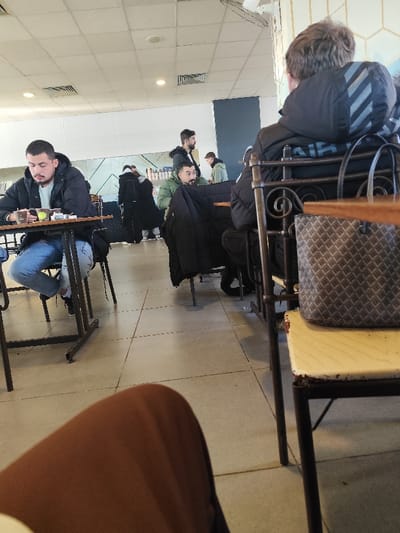 A group of men are in what appears to be a cafe. One man sits to the left, wearing jeans and a dark jacket, seemingly engaged with something on his table. A man in a green jacket sits at another table. A man with a white striped jacket and a black bag is at the far right. The scene is shot from a low angle, including the legs of the person taking the picture.