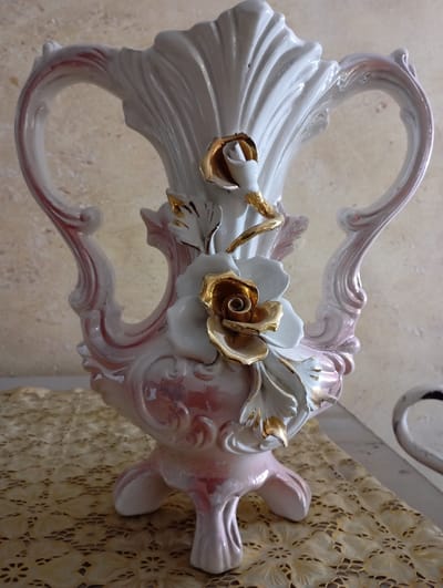 A decorative ceramic vase sits on a textured surface. The vase is primarily white with pink accents and gold detailing, featuring an elaborate design with ornate handles. Two large, white flowers with gold centers adorn the front. The vase has a shell-like textured back and stands on three legs. The background is a soft, neutral color, creating a contrast with the vase's intricate design.