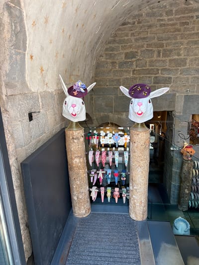The image shows a display of headbands in an arched stone passageway in Bergamo, Italy. Two mannequin heads, designed to resemble white rabbits with open mouths and pink noses, are prominently displayed on wooden logs. Each rabbit head wears a small, decorative purple hat. The head on the left has a hat adorned with blue and purple flowers, while the head on the right is decorated with small, gold bumblebees. A collection of colorful headbands, featuring various patterns and embellishments like flowers, stars, and bows, are displayed on racks behind the rabbit mannequins. The stone walls of the passageway are visible, with painted star motifs on the arched ceiling. The floor appears to be a dark gray mat. The scene suggests a retail or market setting, likely a shop specializing in accessories.