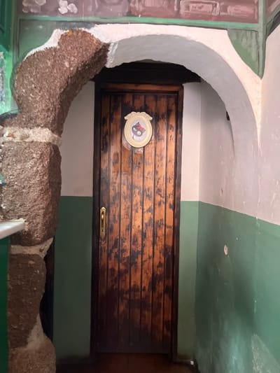The image shows a wooden door in a hallway of a building in Agüimes, Spain, at coordinates 27.907, -15.447. The door is dark brown and has a brass handle on the left side. There is a circular plaque on the door, with a black cat with a red scarf and white whiskers and a white background. The hallway has a light green wall on the right and a white wall on the left. The left wall is partly covered by a large, rounded, brown rock.  The floor is made of tile and there is a small, white counter with a green base on the left side of the doorway.