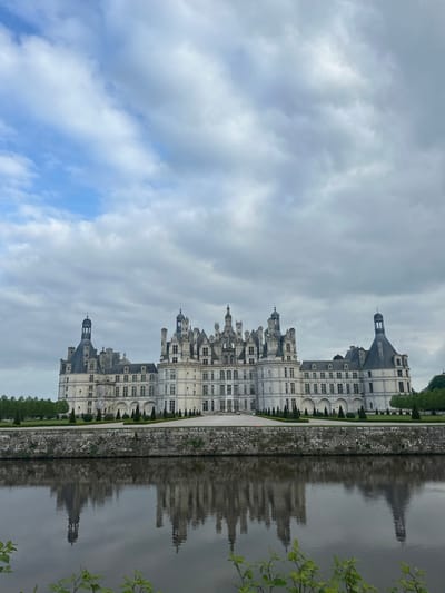 The Château de Chambord, located in the Loire Valley of France (latitude: 47.617, longitude: 1.515), is pictured from a distance in front of a wide moat. The large white castle is made of stone, with a central tower and numerous smaller towers and spires. The castle is symmetrical, with two identical wings extending out from the center. The castle is surrounded by a moat, which is reflected in the water. The water appears calm and still, and the reflection of the castle is clear. The sky is cloudy and overcast. There are no people visible in the picture. A few trees are visible in the foreground and on the banks of the moat, and there are a few trees visible in the background behind the castle.