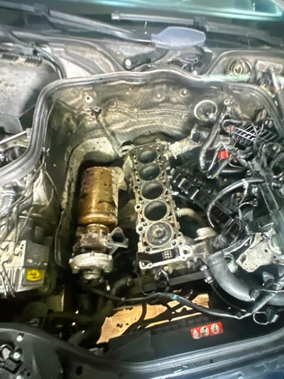 The image displays a highly disassembled car engine bay, indicating major repair or maintenance work. In the center, the engine block is prominently featured with its cylinder head removed, revealing five distinct cylinder bores. Surrounding the block are numerous disconnected hoses, electrical wires, and various engine components, suggesting a complex mechanical operation.
A notable object is a bronze-colored turbocharger, positioned below the exposed engine block, showing signs of thermal wear or accumulation of grime. Adjacent to it, another part of the turbo assembly with a silver-colored housing is visible. Towards the bottom right of the image, an electronic control unit, possibly an ABS module, with a yellow label and associated wiring, can be seen. Parts of what appear to be an intake manifold or other engine covers are visible in the upper left section of the engine bay. A gray, spade-like tool or component is partially visible on the far left.
On the right side of the engine bay, near the vehicle's frame, several red warning stickers are present. These stickers display universal symbols, including a crossed-out hand or object, signifying a prohibition or caution. Other symbols on the stickers are too blurry to be precisely identified, but they are characteristic automotive safety warnings. The overall scene reflects an ongoing intensive automotive repair. No other discernible text is clearly legible in the image.