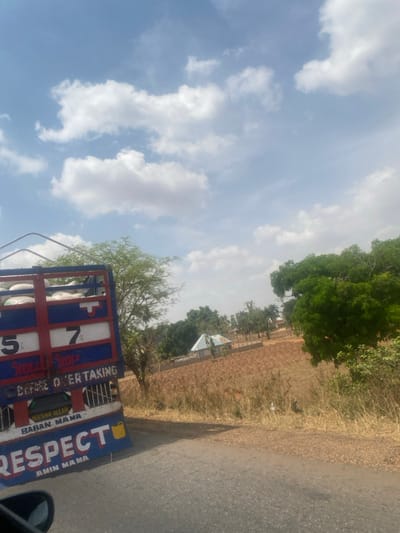 The image captures an outdoor scene in Tataura, Nigeria. A blue and red truck occupies the left foreground, oriented diagonally to the right. Its cargo bed contains multiple white sacks or bundles, secured by metal railings. The side panel of the truck features "5" and "7" in white on red squares, along with painted text including "STOP", "BEFORE OVERTAKING", "MASHA ALLAH", "BABAN MAMA", "RESPECT", and "AMIN MAMA", accompanied by a yellow jerrycan symbol.
An asphalt road runs alongside the truck on its right. The lower-left corner shows a partial view of a dark vehicle mirror, indicating the photograph was taken from inside a vehicle. To the right of the road, a dry, reddish-brown field extends into the mid-ground. Scattered trees, both green and brown, punctuate the landscape. A single-story building with a light-colored, sloped roof, consistent with corrugated metal, is visible in the mid-ground, partially enclosed by a fence. The sky is light blue with numerous scattered white cumulus clouds. The scene is brightly illuminated by natural daylight, casting shadows to the right. No individuals are visible, and no specific actions or interactions are depicted.
