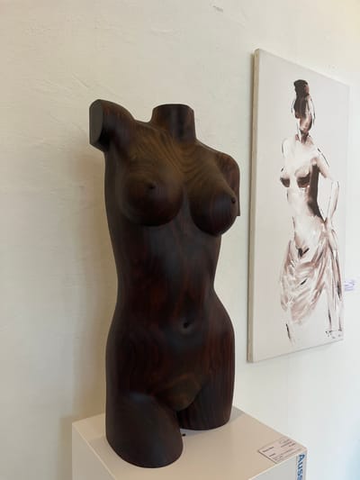 A wooden torso sculpture of a woman's body dominates the foreground. Dark brown and polished, it rests on a white display. Above the sculpture hangs a canvas painting featuring abstract, brown and white brushstrokes, possibly depicting a reclining figure. The background is a plain white wall, creating a minimalist art display.