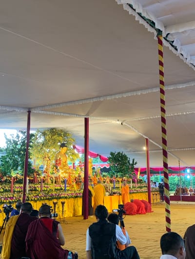 The scene is a large outdoor area with a white tent covered in ruffles, in the village of Mungkid, Indonesia (latitude: -7.605, longitude: 110.23). The tent is supported by poles with red and yellow striped fabric. There is a large golden statue of Buddha in the center, behind a decorative altar. The altar is adorned with flowers, candles, and other offerings. There are several people dressed in orange robes standing near the altar. A group of people are seated in the foreground, watching the scene unfold. Some of these people are holding cameras. The ground is covered in sand. The image was likely taken at an outdoor Buddhist ceremony.