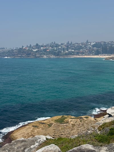 An aerial view depicts a coastline with buildings nestled along the shore, juxtaposed with a vast expanse of blue ocean. The water transitions from a lighter turquoise to a deeper indigo. Rocky cliffs covered in vegetation rise sharply, where white waves crash at the base. The sky is a hazy pale blue, creating a serene yet dramatic coastal landscape.