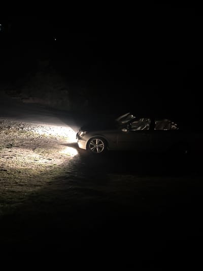 A car has crashed on a dark road, illuminating a mossy embankment in Sveti Konstantin, Bulgaria. The vehicle is upright, with its headlights on.